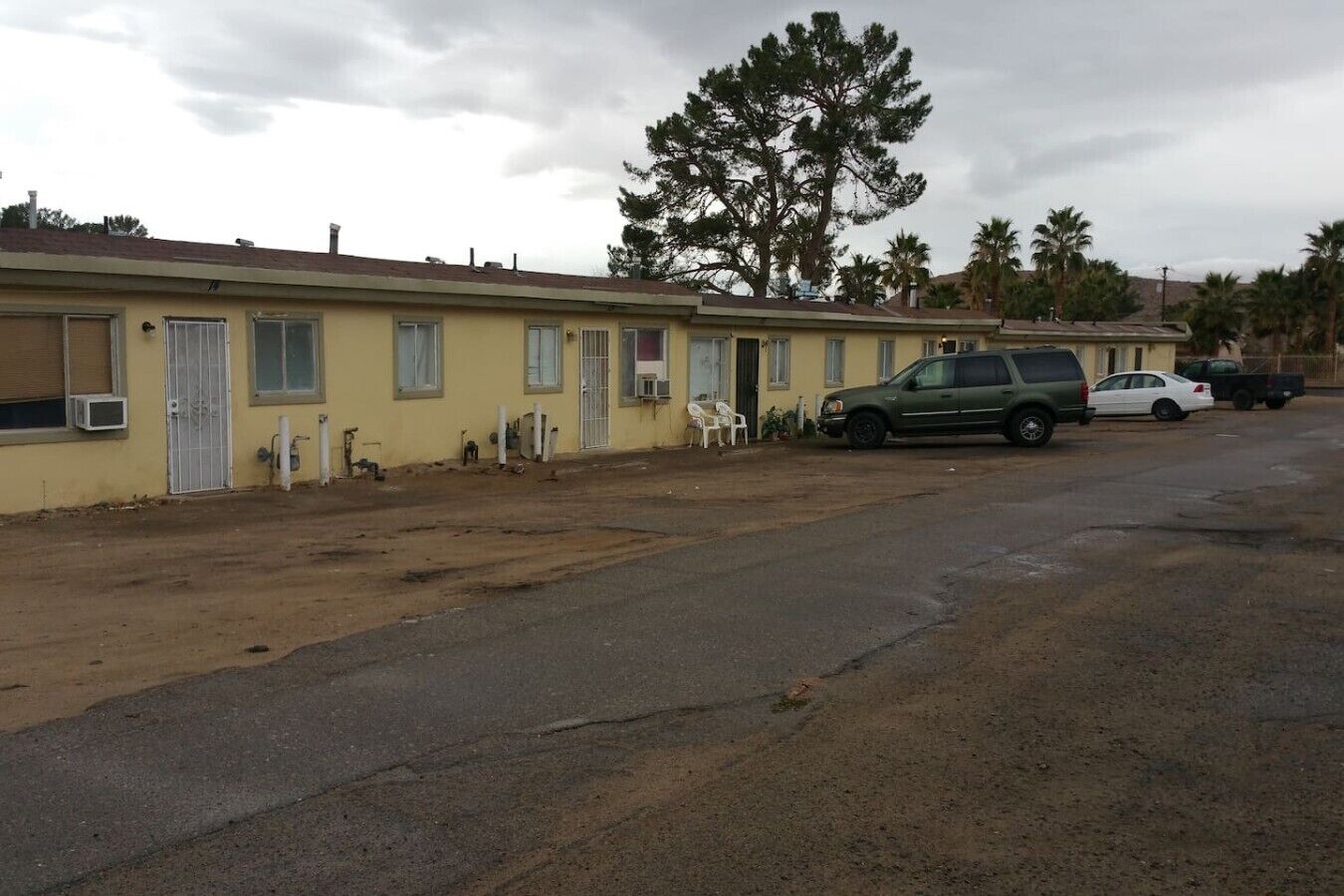 Eine Reihe gelber Gebäude mit unbefestigtem Grund, parkenden Autos und einem großen Baum.