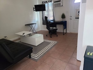 Una sala de estar con un sofá negro, una mesa de centro blanca, una silla de oficina negra, un escritorio y un televisor montado en la pared.