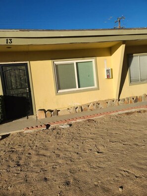A building with a numbered address, a black door, a window with blinds, and a paved area in front.