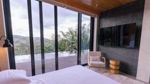 Une chambre dotée d’un grand lit, d’une chaise, d’un téléviseur fixé à un mur texturé, et offrant une vue sur un balcon avec une terrasse en bois et de la verdure.