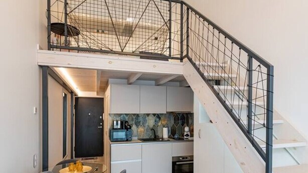 A modern apartment with a grey sofa, a kitchen with white cabinets, and a staircase with a metal railing. - Sunshine Loft by Polo Apartments (Kaunas)