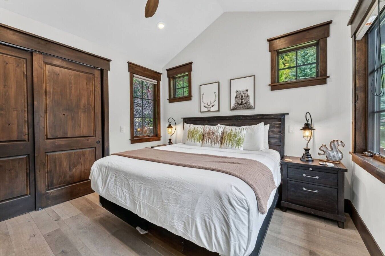 A bedroom with a large bed, wooden wardrobe, two bedside tables with lamps, and a window with a view of greenery.
