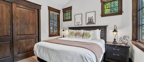 A bedroom with a large bed, wooden wardrobe, two bedside tables with lamps, and a window with a view of greenery.