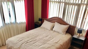 Un lit avec une couette beige, deux oreillers, une tête de lit en bois, deux tables de chevet avec des lampes et une fenêtre avec des rideaux rouges.