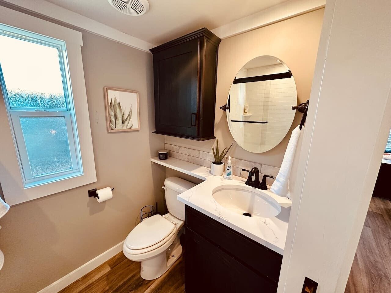 A bathroom with a toilet, sink, dark cabinets, a mirror, and a window.