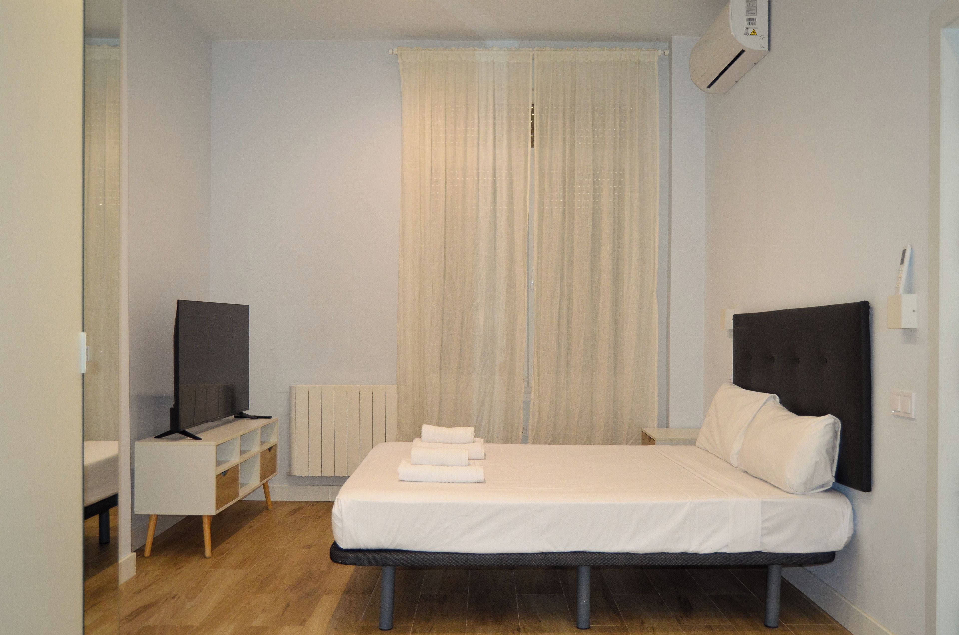 Una cama individual con cabecera, una mesita de noche, un televisor y una ventana con cortinas.