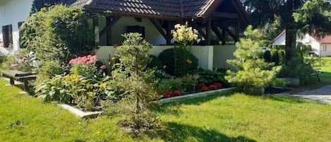 Una casa con techo inclinado, un jardín con diversas plantas y flores, y una terraza de madera.
