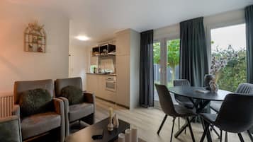 A modern living room with a sofa, a dining table with chairs, a kitchen area, and a view of greenery outside.