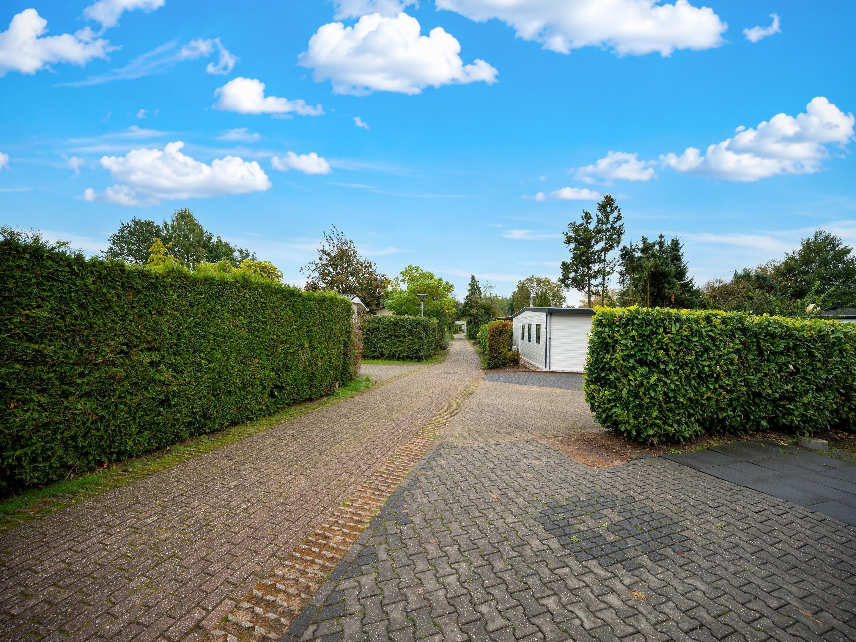 A paved pathway leading to a white building with a dark roof, surrounded by neatly trimmed hedges and trees.