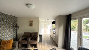 A modern living room with a sofa, two armchairs, a small table, a decorative mirror, and a view of a kitchen and outdoor area. - Chalet 56 in Stegeren Near the Heathlands (Stegeren)