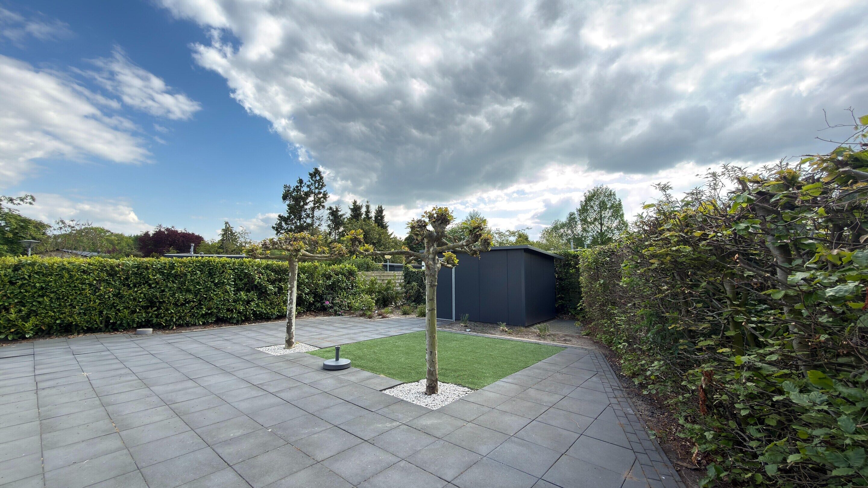 A paved patio with a small lawn area, a shed, and a hedge.