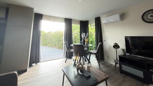 A modern living room with a flat-screen TV, a dining area with chairs, a coffee table, and a large window with curtains. - Chalet 56 in Stegeren Near the Heathlands (Stegeren)