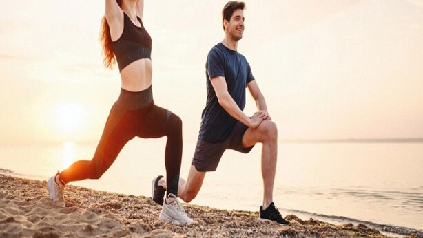 Due persone che eseguono esercizi di stretching su una spiaggia con il tramonto sullo sfondo.