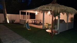 A covered outdoor area with a table and chairs, a slide, and a thatched roof structure. - Large Holiday Homes With Beautiful Gardens (Rewal)