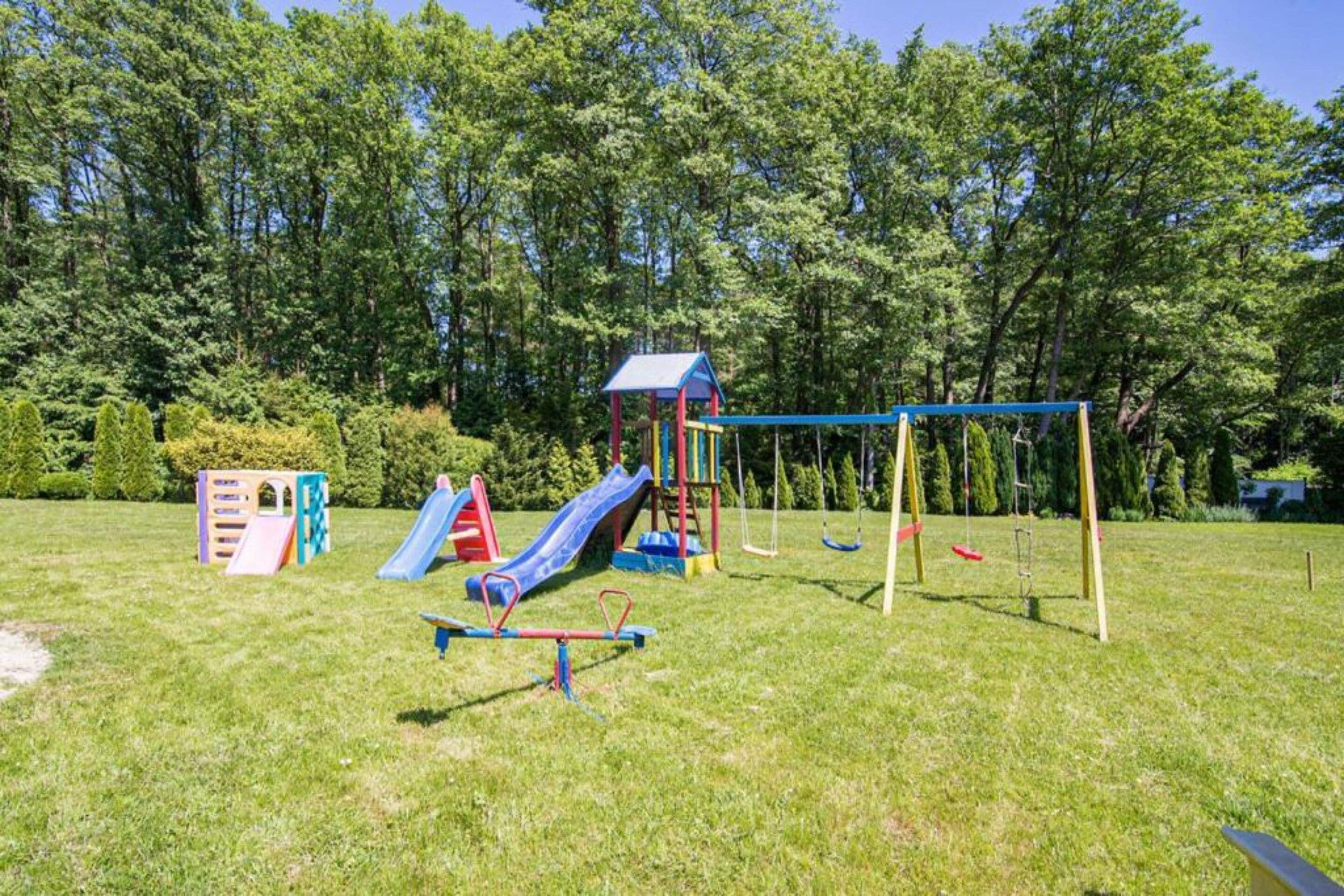 A playground with slides and swings in a grassy area surrounded by trees.