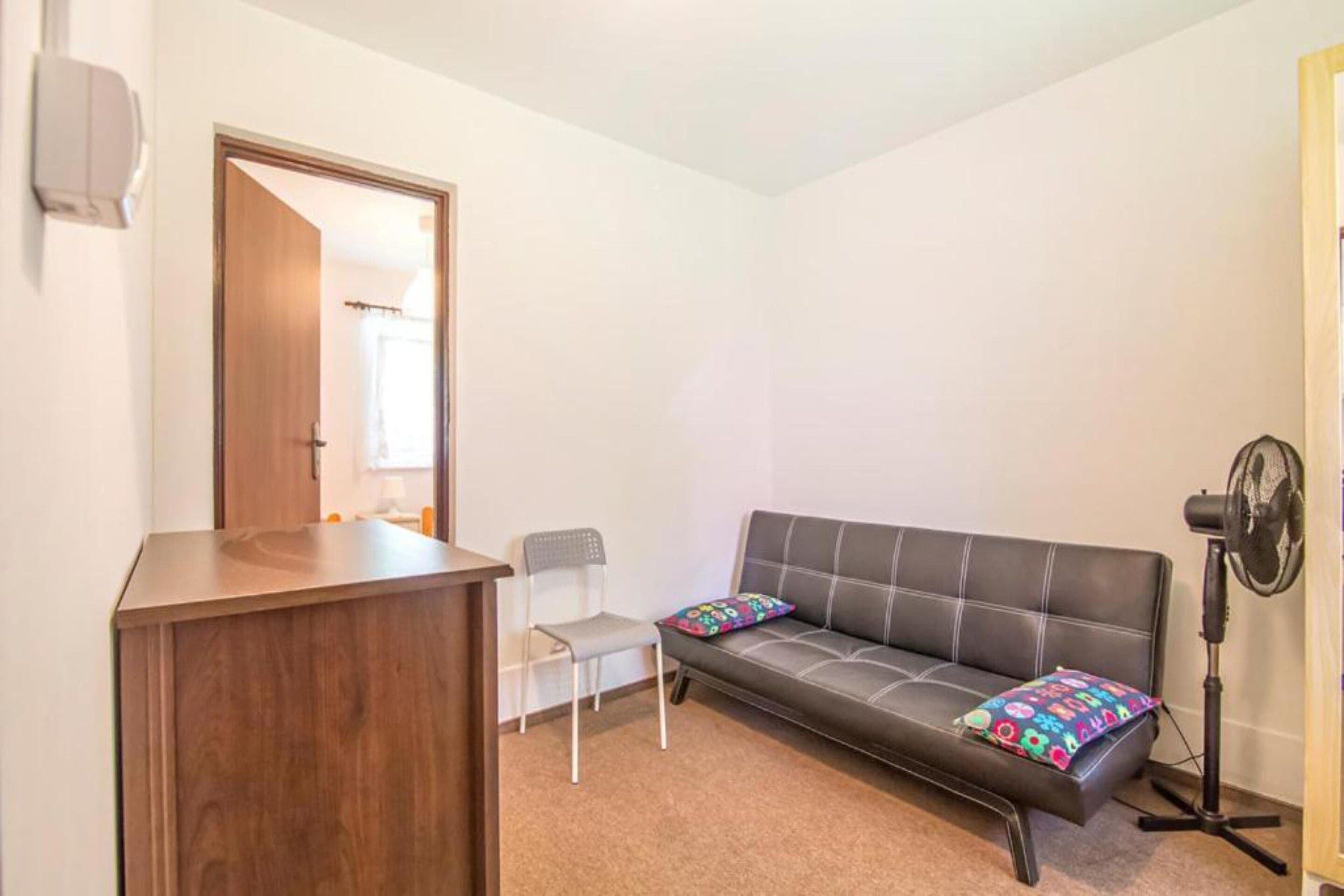A small, furnished room with a wooden desk, a grey sofa with colorful cushions, a single white chair, and a standing fan.