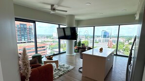 A modern living room with a large TV, a sofa, a coffee table, and a view of the cityscape through large windows.
