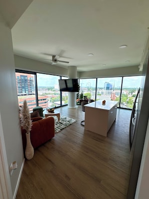 A modern living room with a large TV, a sofa, a coffee table, and a view of the cityscape through large windows. - Luxury Raleigh Condo w/ Panoramic City Sunset Views (Raleigh)