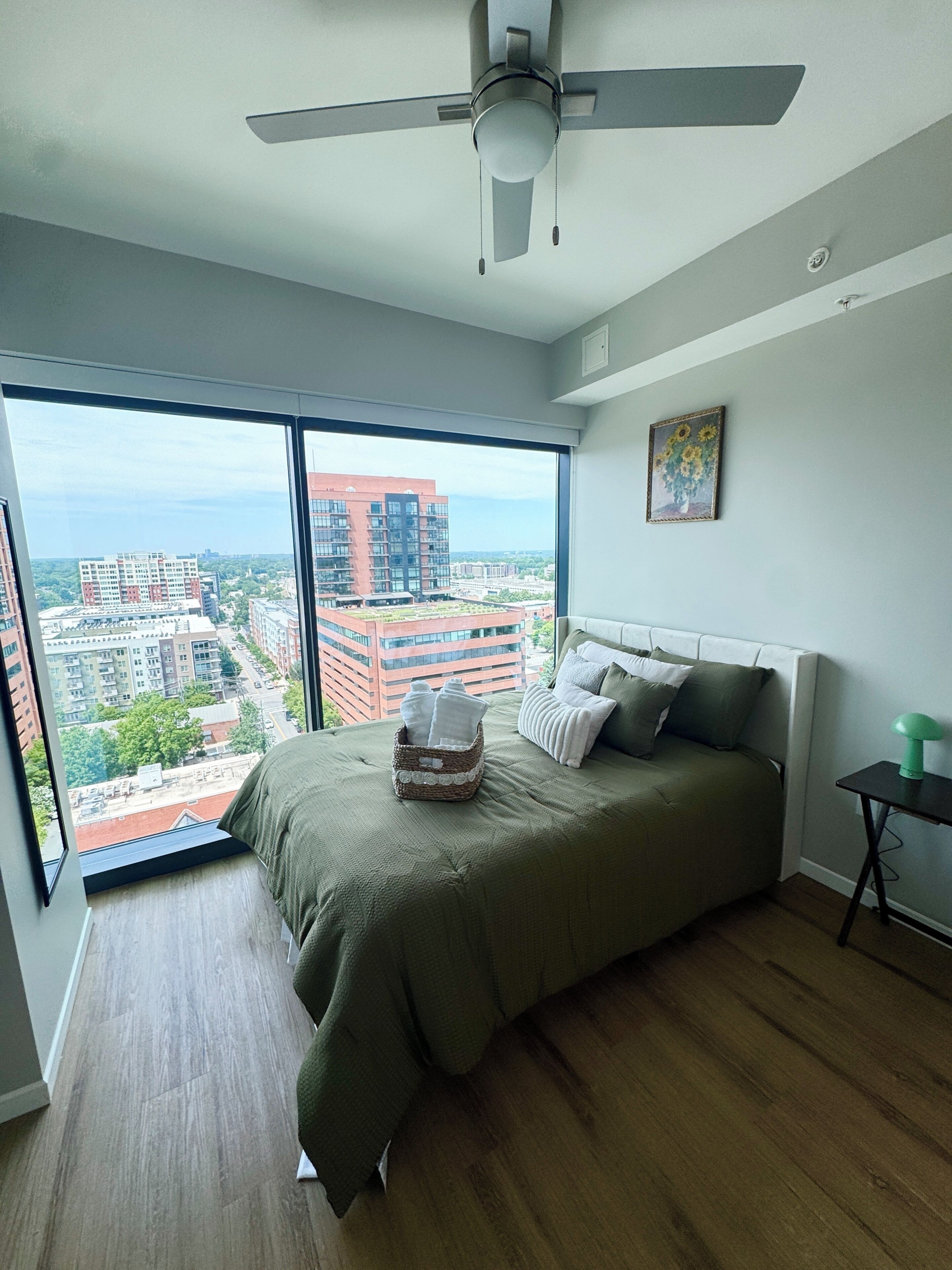 A bedroom with a large window, a ceiling fan, a bed with pillows, a bedside table, and a painting on the wall.
