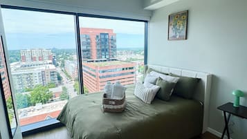 A bedroom with a large window, a ceiling fan, a bed with pillows, a bedside table, and a painting on the wall.