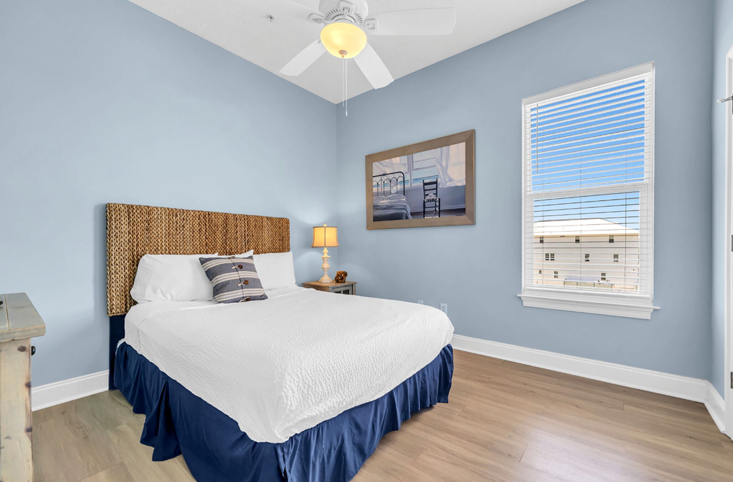 Ein Schlafzimmer mit Bett, Nachttisch, Lampe, Deckenventilator und Fenster mit Jalousien.