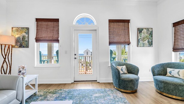 A room with a white door, two windows with brown blinds, a blue armchair, a white sofa, and a patterned rug. - Vista del Mar II - Coastal Calm PCB 301 (Panama City Beach)