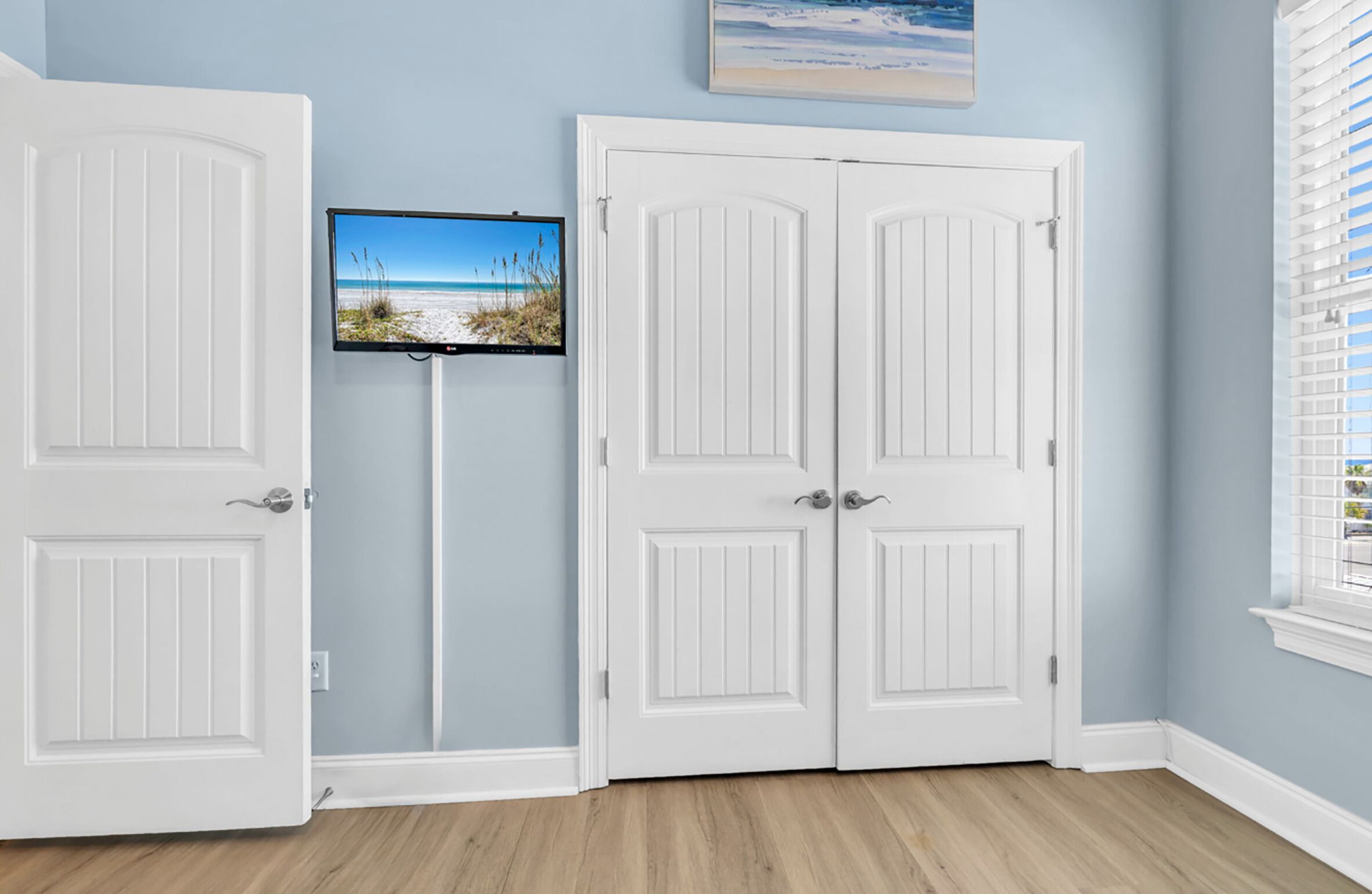 Una habitación con dos puertas blancas, un televisor montado y una ventana con persianas.