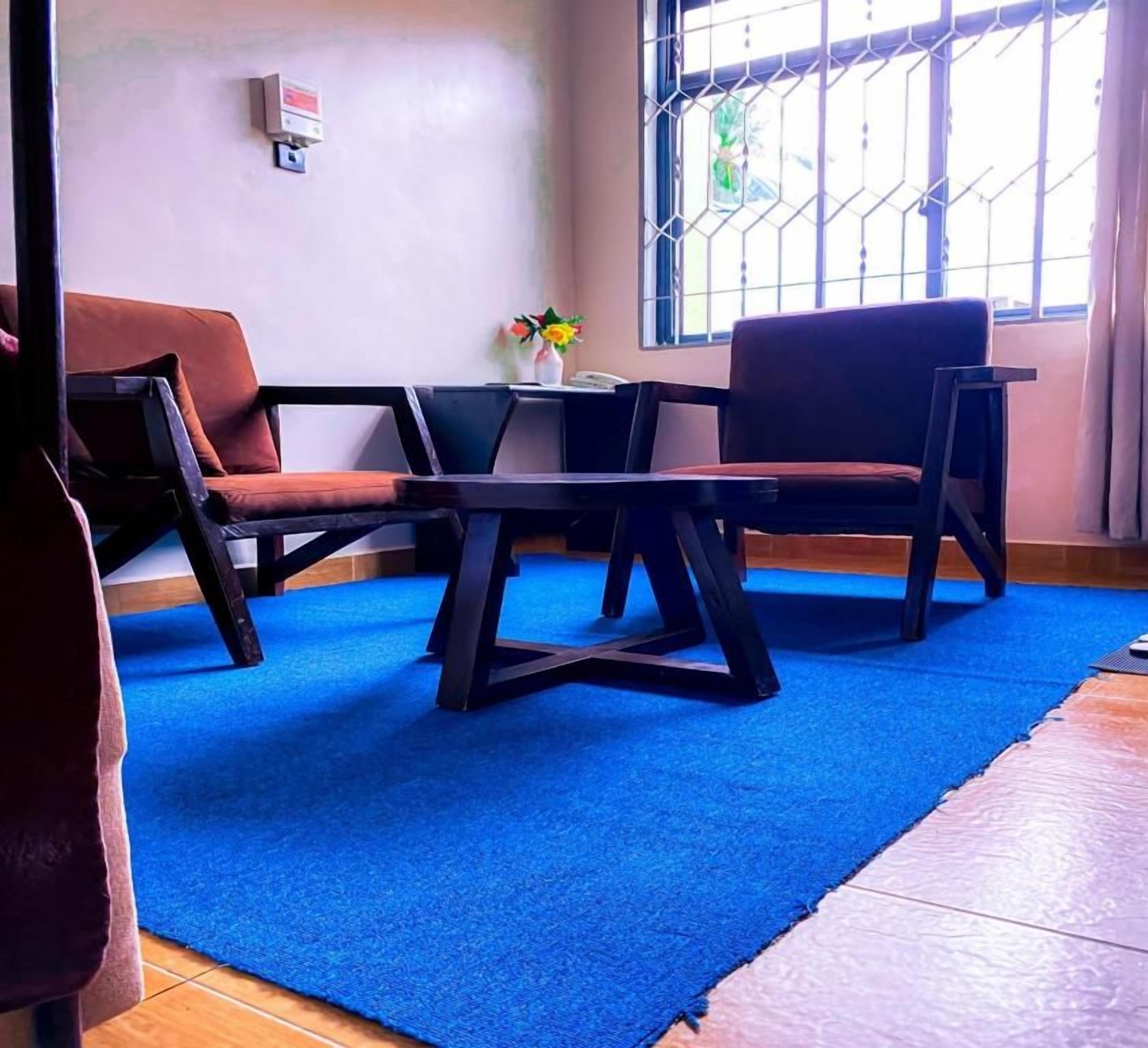 Una habitación con alfombra azul, dos sofás marrones, una mesita con un florero y una ventana con barrotes.