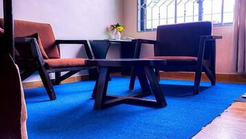 Una habitación con alfombra azul, dos sofás marrones, una mesita con un florero y una ventana con barrotes.