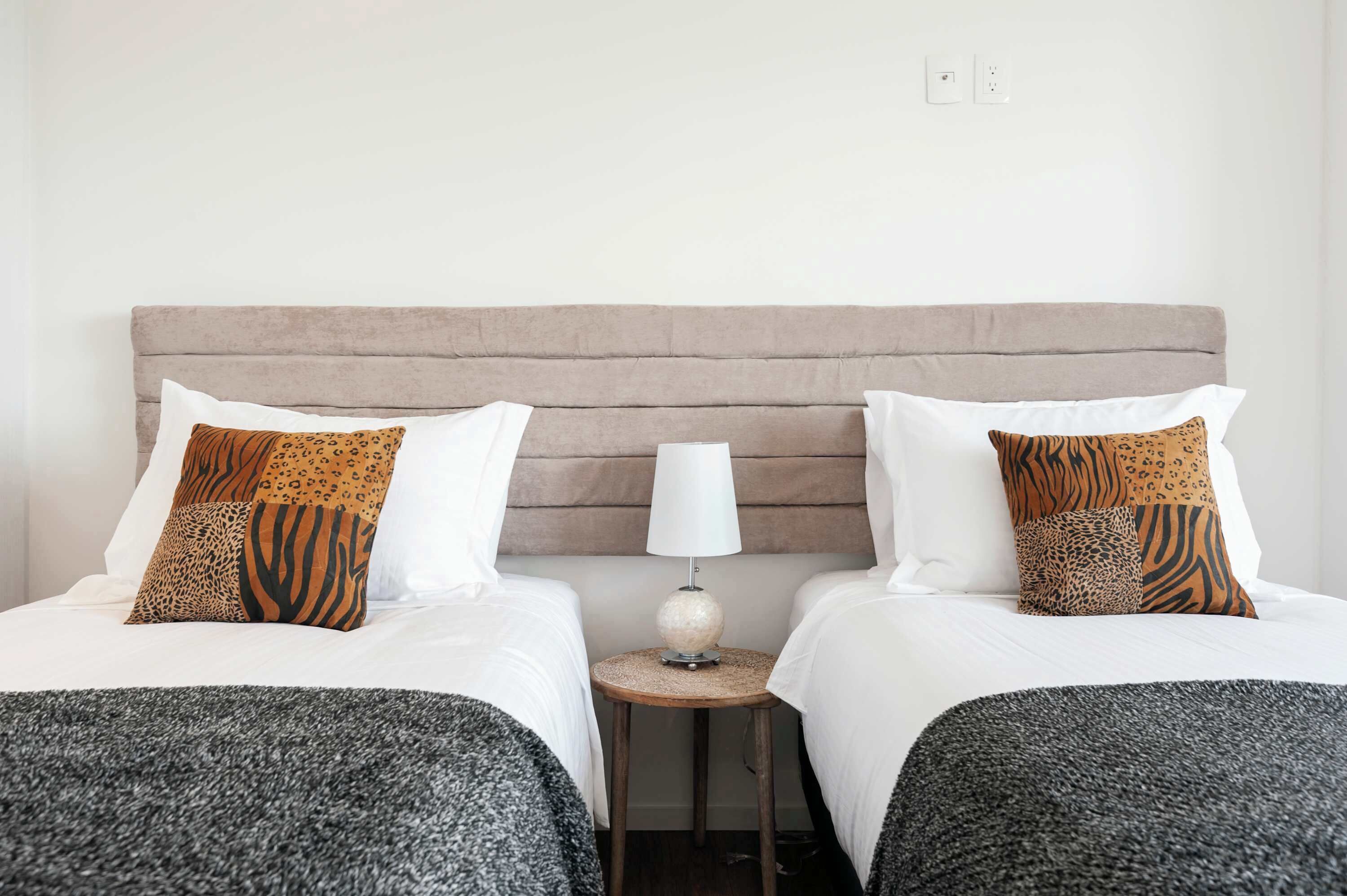 Habitación de hotel con dos camas, cada una con ropa blanca y almohadas estampadas, una mesita redonda con una lámpara y un cabecero de color neutro.