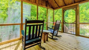 Una terraza cubierta de madera con vistas a un bosque, dos mecedoras y una mesa.