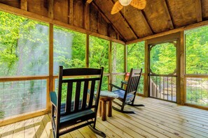 A covered wooden deck with a view of a forest, two rocking chairs, and a table. - 15 Minutes from Skyland Ranch. Brand New Cozy Cabin. Huckleberry Cabin (Sevierville)