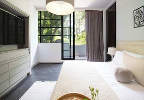 Ein modernes Schlafzimmer mit einem großen Bett, einem Fernseher, einem Fenster mit Vorhängen und Blick auf Grünflächen.