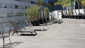 Una terraza en la azotea con tumbonas, palmeras y un edificio moderno al fondo.