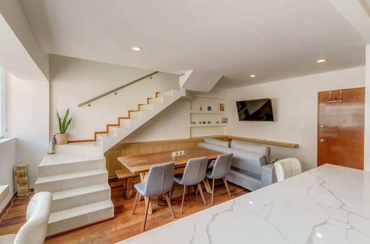 Een modern interieur met een trap, een eethoek met stoelen, een bank en een aan de muur bevestigde televisie.