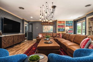 A living room with a sectional sofa, a flat-screen TV, a chandelier, and a bookshelf. - Cedar Hall - A Charming Vintage Estate (Lebanon)