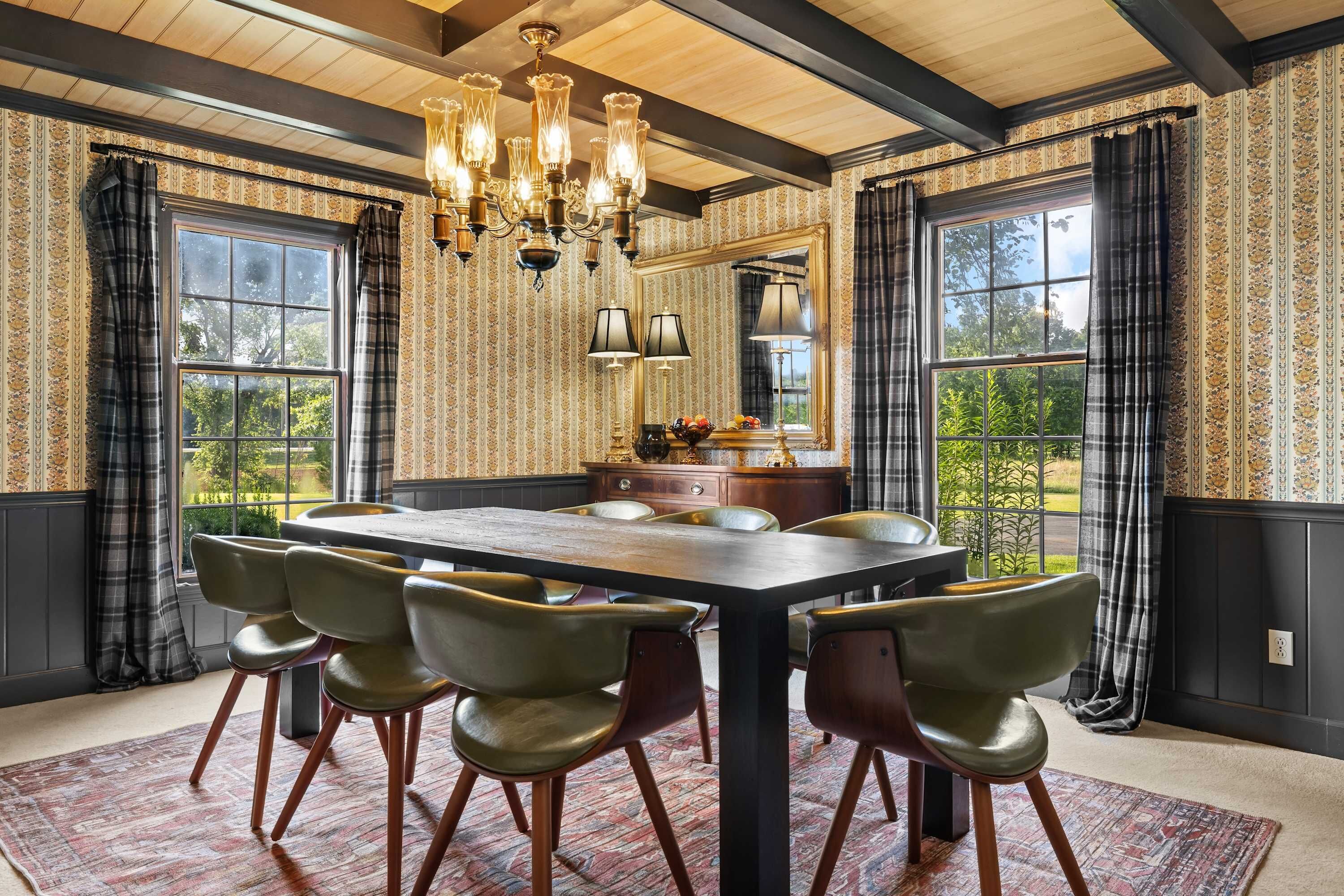 A dining room with a large table, six chairs, a chandelier, and large windows with curtains.