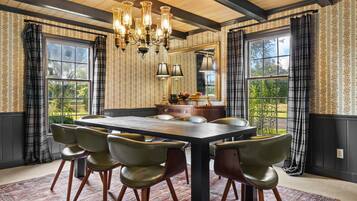 A dining room with a large table, six chairs, a chandelier, and large windows with curtains.