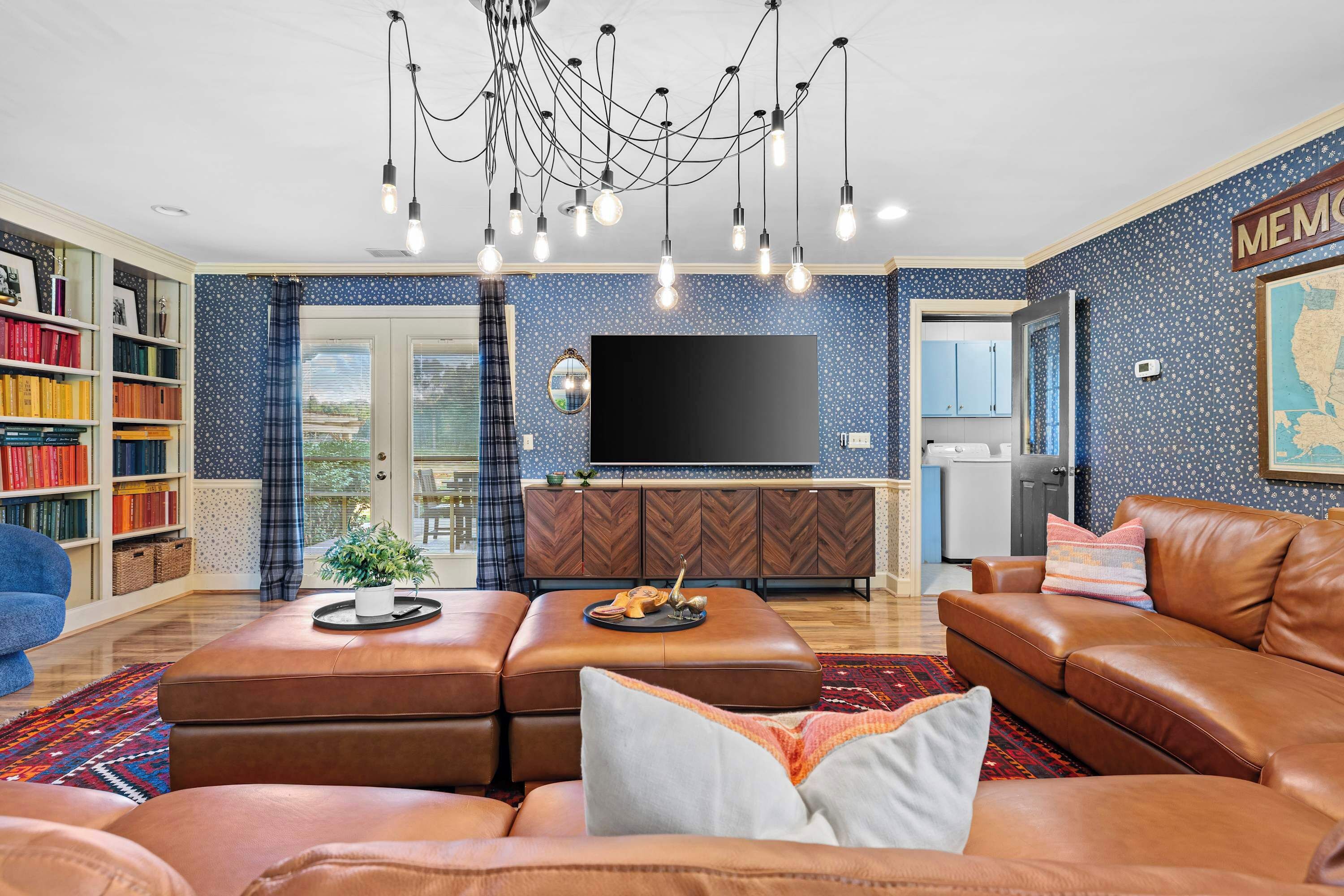 A living room with a brown leather sofa, a coffee table, a bookshelf, a flat-screen TV, and a chandelier.