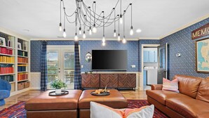 A living room with a brown leather sofa, a coffee table, a bookshelf, a flat-screen TV, and a chandelier. - Cedar Hall - A Charming Vintage Estate (Lebanon)