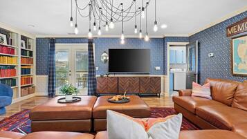 A living room with a brown leather sofa, a coffee table, a bookshelf, a flat-screen TV, and a chandelier.