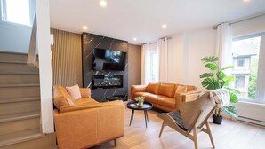 Een moderne woonkamer met een bank, een fauteuil, een televisie, een open haard en een groot raam.