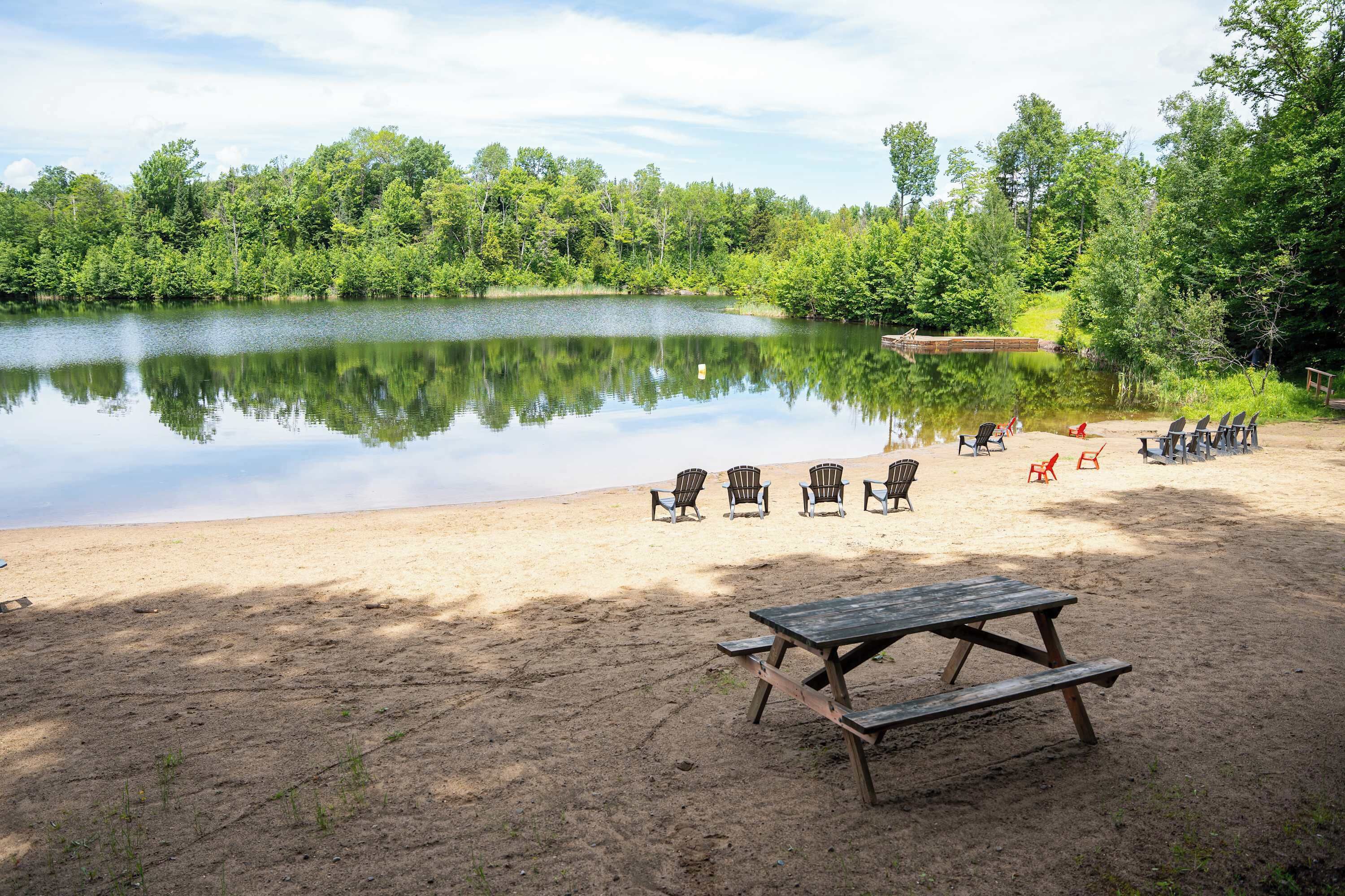 Une plage de sable bordée d’un lac paisible, entourée d’arbres, et comprenant une table de pique-nique et des chaises.