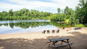Een zandstrand met een rustig meer, omringd door bomen, met een picknicktafel en stoelen.