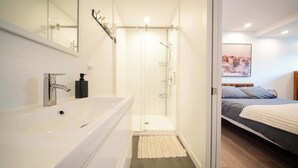 Een moderne badkamer met een groot wit wastafelmeubel, een douchecabine met glazen wanden en een bed met een grijs-witte dekbedovertrek.