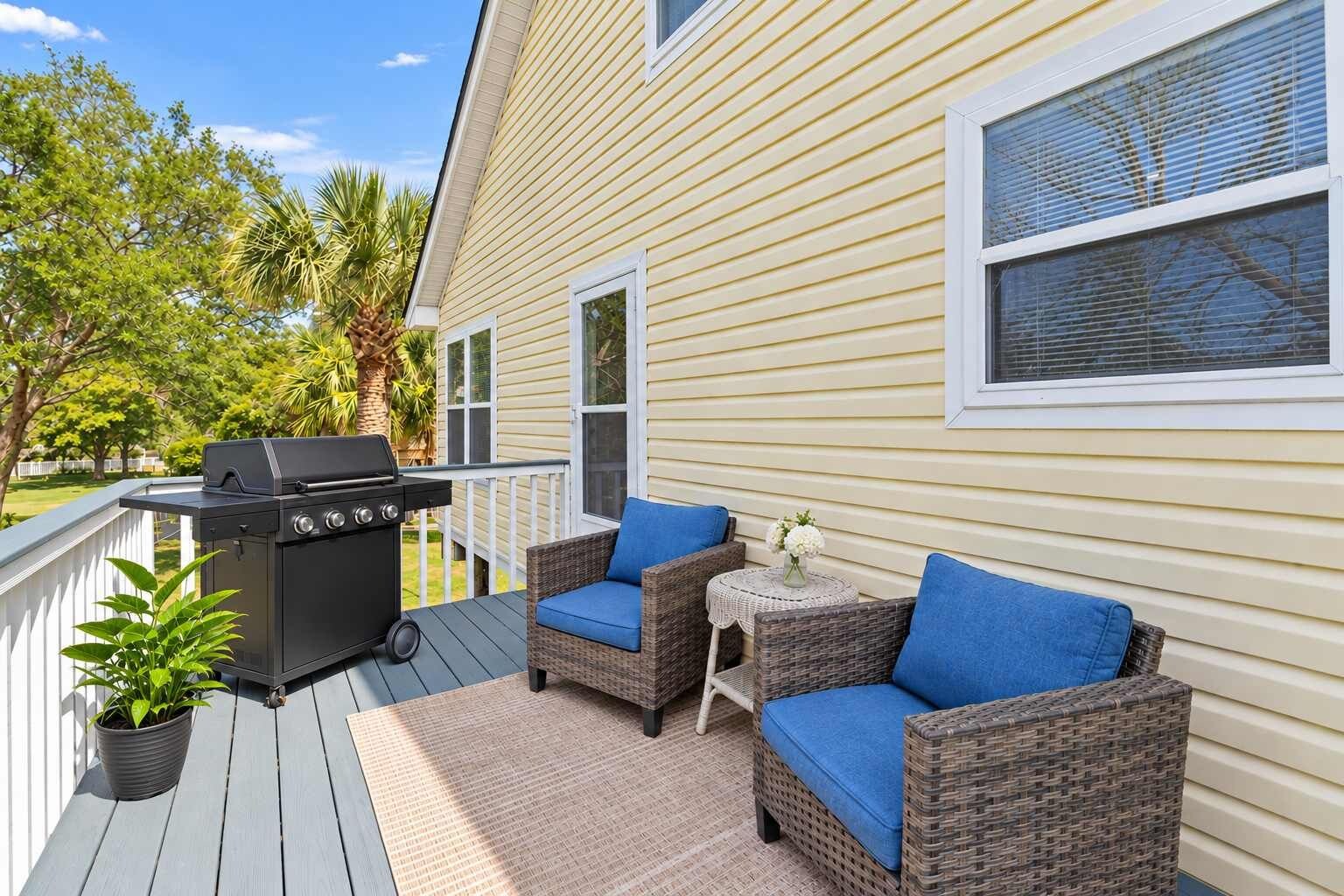 A deck with a grill, two wicker chairs with blue cushions, and a small table with a vase of flowers.