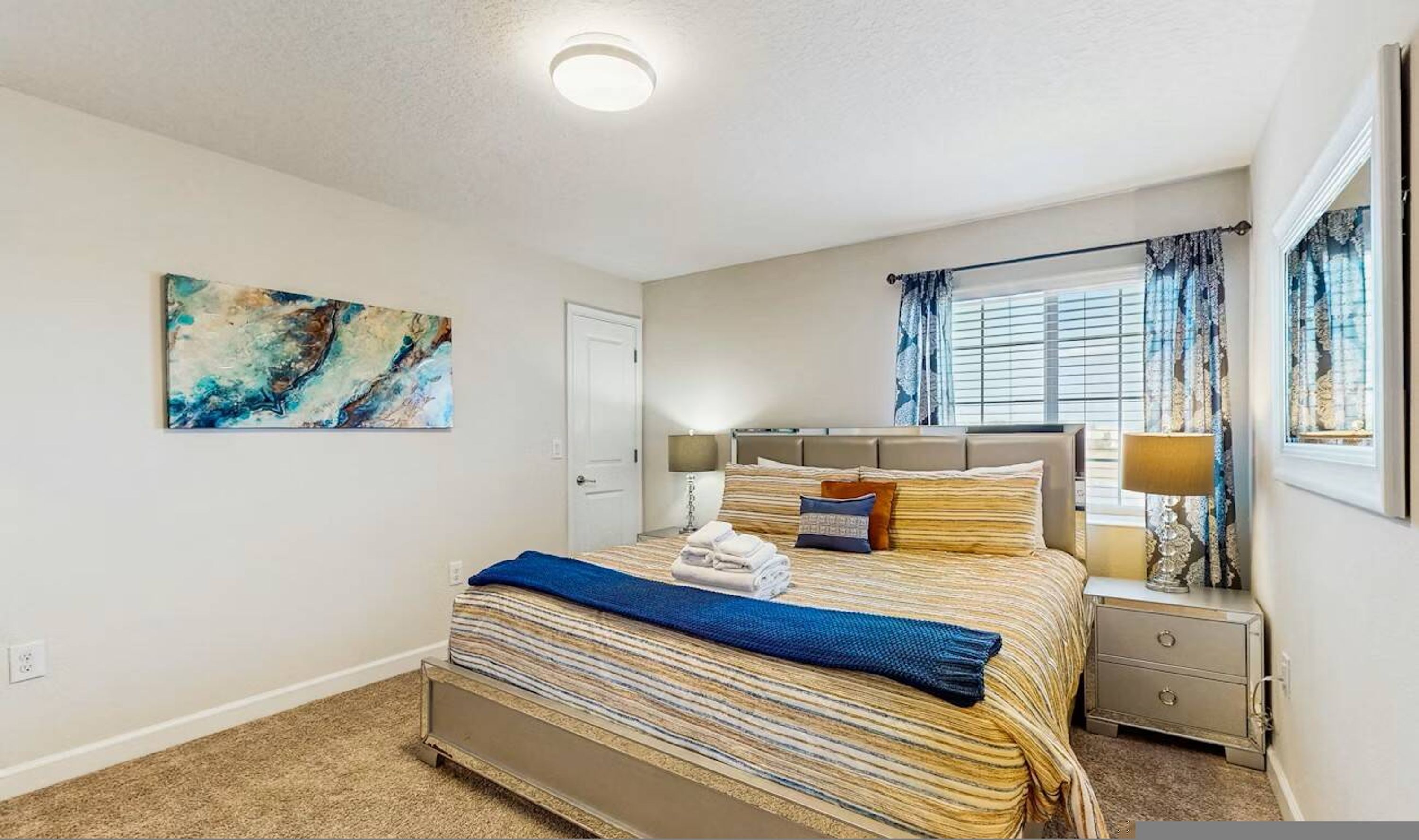 Quarto com cama, mesas de cabeceira, cortinas e um quadro na parede.