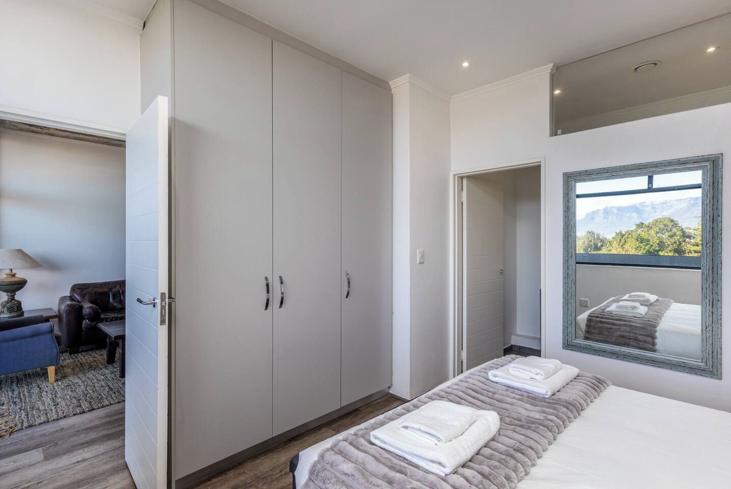 A bedroom with a large wardrobe, a bed with towels, a window with a view, and a small seating area with a lamp.