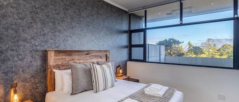 A modern bedroom with a large bed, wooden headboard, and a view of a mountain landscape through a large window.