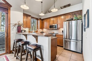 A kitchen with wooden cabinets, a stainless steel refrigerator, a microwave, and a large island with stools. - Breck Basecamp | Deck + Hot Tub + Walk Everywhere (Breckenridge)
